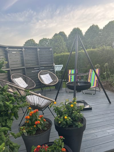 The image depicts a backyard patio area with a wooden deck. In the foreground, there are two large black planters containing flowering plants, one with orange and red blooms, and another with yellow flowers. Behind the planters, several round, woven chairs with white striped cushions are arranged. To the right of the chairs, a black tripod grill stand with a fire pit is set up, and two striped folding chairs are placed behind it. A dark wooden fence runs along the back of the patio, and beyond that, a line of green trees and a hedge are visible. The sky above is partly cloudy and overcast. The scene appears to be in a residential area, likely in Sandefjord, Norway, given the information provided.