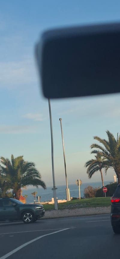 The image depicts a roadside scene in Harhoura, Morocco, captured from inside a vehicle, with a dark, blurred object (likely a dashboard or rearview mirror) partially obscuring the upper frame. The scene presents a coastal environment under a clear, light blue sky.
In the foreground, an asphalt road surface features a white solid line on the left and a double solid white line further right, indicating traffic lanes. A dark green SUV, possibly a Dacia Duster, is positioned on the left side of the road, facing left, with its front quarter and a distinctive silver and black wheel visible. Its daytime running lights are illuminated. On the far right, the rear quarter and illuminated red taillight of a dark-colored SUV are visible, facing away from the viewer.
Between the road and the ocean, a raised concrete or paved barrier supports a grassy median strip adorned with several mature palm trees. Multiple tall, slender, metallic poles (likely streetlights or flagpoles) are prominently erected within this strip. A circular black-and-white regulatory sign and a circular red 'no entry' sign are visible among the foliage on the right side of the median.
The mid-ground reveals the calm, blue expanse of the Atlantic Ocean, with gentle waves breaking near the shore. Small, white, indistinct structures, potentially boats or coastal features, are visible closer to the waterline. The horizon line where the sky meets the ocean is clearly defined. The overall lighting suggests either early morning or late afternoon conditions. No individuals are discernible within the frame.