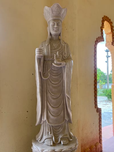 A full-length, pale beige marble statue of a crowned deity stands against a plain, light yellow-beige wall in Đăk Mar, Vietnam. The statue wears a detailed headdress and a necklace, holding an upright cylindrical object in its raised right hand and cradling a flame-shaped object in its left hand. Its flowing robes exhibit subtle linear patterns. The statue rests on a carved lotus-petal base. To the right, an ornate archway with a textured, reddish-brown border frames an outdoor view. This view reveals a paved ground, green foliage, and a utility pole against a bright, clear sky.