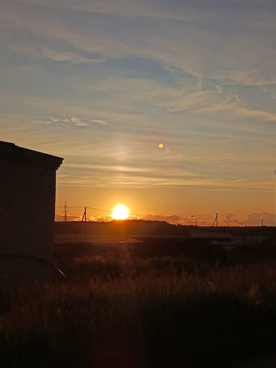 This is a vertical, eye-level, full-color photograph of a sunset over a rural or industrial area, possibly Kauchuk Stantsiya, Russia, given the context provided. The sun, a bright orange disc, is low on the horizon, partially obscured by a line of trees and some clouds. A smaller, pale disc, possibly the moon or a lens flare, is visible higher in the sky. The sky transitions from a pale blue at the top to a warm orange near the horizon, with scattered wispy white clouds.

In the foreground, the left side of the image is dominated by the dark silhouette of the side of a brick building, with its roofline angled downwards towards the right. A field of dry, tall grass, mostly in shadow but catching some of the sunset's orange light, occupies the lower portion of the frame.

In the middle ground, beyond the field, a flat expanse is dotted with indistinct structures and silhouettes of trees. Power lines and utility poles are visible, with one tall, lattice-like tower standing out. The setting sun casts a warm glow over this landscape.

The overall impression is one of a peaceful, end-of-day scene in a sparsely populated or developing area. There are no people visible in the image.