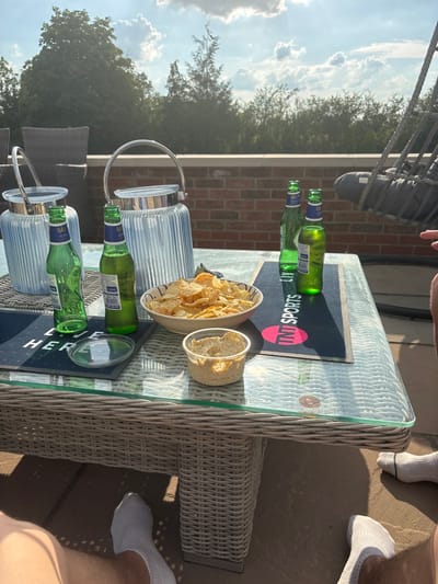 A wicker table outside holds an array of snacks and drinks. A bowl overflows with potato chips, accompanied by a smaller container of dip. Several green beer bottles lay empty on a TNT Sports mat, joined by two glass pitchers. A glimpse of a brick wall and trees is visible in the background. The table rests on a stone patio, where feet in white socks can be seen at the table's edges.