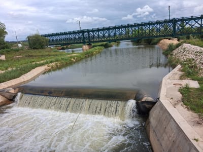 The image shows a weir in a canal in Montemor-o-Velho, Portugal. The water flows over the weir, creating a small waterfall effect with white water at the base. The canal is concrete-lined on either side. A green steel truss bridge spans the canal in the background. Vegetation grows along the banks of the canal. The sky is overcast.