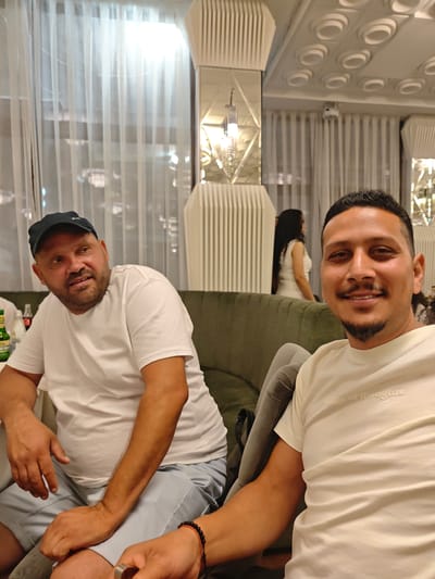 Two men are seated in a restaurant, likely in the city of Peshtera, Bulgaria, as indicated by the presence of a bottle of Zagorka beer. The man on the left is wearing a white t-shirt and a black baseball cap. He has a beard and appears to be speaking. The man on the right is closer to the camera, also wearing a white t-shirt with subtle text that reads "edge of thoughts". He has a goatee and is smiling directly at the camera. In the background, another woman with long dark hair, wearing a white dress, is visible, blurred by the shallow depth of field. The restaurant interior features decorative ceiling elements and large windows with sheer white curtains. The seating appears to be a dark green couch.