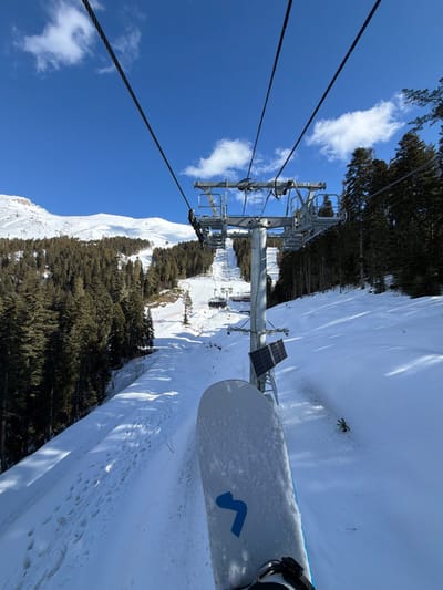 The image provides a first-person perspective from a ski lift chair at a ski resort in Lunnaya Polyana, Russia, during daytime under clear skies. In the immediate foreground, the top surface of a white snowboard, partially covered in snow and featuring a blue 'S' or '3' shaped logo near the binding, is visible. The snowboard is attached to a boot, indicating the observer's position on the lift. Directly ahead and slightly to the right stands a tall metal ski lift tower, identified by the number 14, from which multiple parallel cables extend. Two small, dark rectangular solar panels are affixed to the side of this tower. Below, a snow-covered ski slope descends, bordered on both sides by dense coniferous trees. Ski tracks and footprints are visible on the slope. Further down the slope, additional ski lift chairs, some occupied by indistinct figures, are observed ascending towards another ski lift tower. A section of red safety netting is also visible further down the slope. In the background, extensive snow-covered mountains are visible against a bright blue sky with a few scattered white clouds.