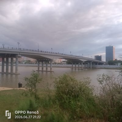 The image shows a wide, modern concrete bridge spanning a calm, brownish river in Atyrau, Kazakhstan. The bridge, supported by multiple large concrete pillars, has a railing along its top and streetlights spaced intermittently. People are visible walking on the bridge. On the far bank, there are buildings, including a tall, slender glass skyscraper with horizontal window bands and a darker, boxier building behind it. The sky is overcast with gray clouds. In the foreground, the bank of the river is covered with green grass and dense bushes. On the near bank, two figures, possibly men, are standing near the water's edge. A timestamp in the lower left corner reads "OPPO Reno3 2026.06.27 21:22". The scene appears to be set in the evening.