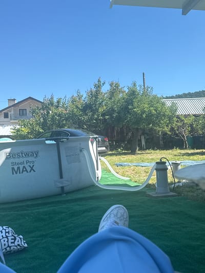 The image shows a Bestway Steel Pro MAX swimming pool set up on a green artificial turf in a residential backyard. A filtration pump is connected to the pool via white hoses. In the background, there is a black car parked in front of a wooden structure and a house. Trees and a metal-roofed building are also visible. The sky is clear and blue. The scene appears to be located in the city of Suvorovo, Bulgaria, based on the context provided. A person's legs and feet, wearing white sneakers and blue clothing, are visible in the foreground, suggesting they are relaxing near the pool.