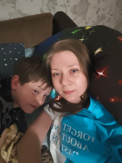 In the image, a young boy and a woman are lying down, possibly on a couch or bed, in what appears to be a domestic setting. The boy is positioned to the left, with his head resting on a pillow with a starry pattern. He has brown hair and is looking towards the camera with a slight smile. The woman is to the right, also looking at the camera, with long brown hair and blue eyes. She is wearing a blue sweatshirt with white text that reads "FORGET ABOUT PAST" in reverse. The background shows a patterned wallpaper and a dark, star-patterned pillow. There are no visible indications of the specific location being Малая Валяевка, Russia. The overall mood of the image is relaxed and intimate, suggesting a casual moment between family members.