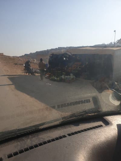 The image, captured from the interior of a vehicle, shows a vibrant roadside scene in Killi, Syria, under a clear, bright sky. The foreground reveals parts of a vehicle's dashboard and windshield, including air vents and reflections, suggesting the viewer is looking out from the passenger or driver's side.
Outside the vehicle, a dusty, unpaved road or path leads to a temporary market stall on the right side of the frame. This stall is sheltered by a light-colored tarp or canopy, beneath which numerous items, predominantly colorful flowers or garlands in shades of orange, white, and green, are displayed. These goods suggest the stall might be selling religious offerings or decorations.
Two people are visible near the stall. One individual, dressed in lighter attire, stands near the displayed goods, seemingly attending the stall. Another person, in darker clothing, is seated on a low stool or bench, next to a small, dark-colored animal, possibly a donkey or mule, which appears to be harnessed or standing alongside a cart. The presence of the animal suggests its use for transport or labor.
The background shows a distant landscape with low-lying hills or buildings under a bright, cloudless sky. The overall environment appears dry and dusty, characteristic of an arid region. The strong sunlight and clear conditions indicate it is daytime. No discernible text is visible in the image. The scene depicts a quiet moment of daily commerce or activity in a rural or semi-urban setting within Killi, Syria.