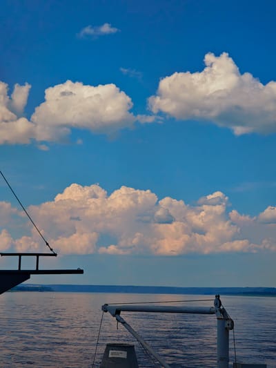 The image depicts a body of water, likely a river, under a partly cloudy sky. The water surface exhibits gentle ripples, reflecting the light of the sky. In the foreground, a portion of a vessel is visible, including metallic structures and rigging. A darker, angled structure on the left appears to be part of the ship's deck or superstructure. To the right, a lighter colored, angled arm with pulleys and cables extends into the frame. A cylindrical object, possibly a storage bin or equipment housing, is situated at the bottom right, bearing some white text. In the mid-ground, the opposite shore is visible as a dark, tree-lined expanse under a hazy sky. The overall scene suggests a calm day on the water, possibly during daylight hours given the brightness of the sky and clouds.
There is no information in the image to suggest the location is the city of Podkamennaya Tunguska, Russia.