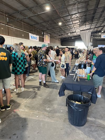 The image shows a large crowd of people in an indoor market, most likely in a warehouse space with a corrugated iron roof, at the Hamilton, Australia location, based on coordinates (-27.44, 153.072).  The market is set up with several tables, a trash can, and various vendors selling food and drinks. People are browsing and purchasing items. Some are interacting with vendors, while others are engaged in conversation.  There is a sign overhead with "SAKE" written in blue letters.  The scene is illuminated by overhead lights.  The floor is a rough concrete surface.  A large group of people is congregated near a table with a black tablecloth and a black trash can.  To the right of the trash can, several people are interacting with a vendor behind the table.  To the left, a man in a green Hawaiian shirt is talking to a woman wearing brown pants.  The vendor is holding a jar of something in his left hand and appears to be speaking to a woman wearing a black and white checkered shirt, who is looking off-camera.  Many people are milling about the venue, some walking, some talking.  The overall atmosphere is vibrant and lively.