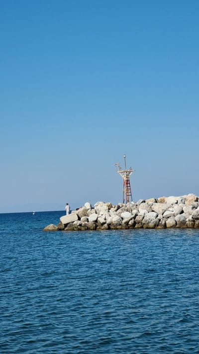 A low-angle, vertical, full shot captures a bright, clear day at the port of Limani Litochorou, Greece. The foreground is dominated by the deep blue, slightly rippled surface of the sea, with a rocky breakwater stretching from the right to the mid-left of the frame. A solitary figure, dressed in white, stands on the breakwater, facing away from the camera and towards the sea. To the right of the figure, on the breakwater, stands a navigational aid structure with a ladder. The background is a vast expanse of clear, pale blue sky. In the distance, a small white boat is visible on the water to the left of the breakwater. The overall impression is one of calm and tranquility.