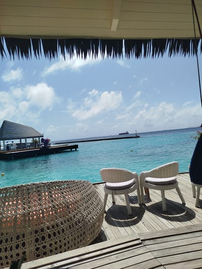 The image depicts a serene scene at Gulhi Falhu, Maldives, featuring a picturesque view of the turquoise ocean. In the foreground, a woven lounge chair is positioned on a wooden deck. Further back, two white wicker chairs with light grey cushions are arranged on the same deck, casting distinct shadows. Beyond the deck, a wooden jetty extends into the calm water, supporting a thatched-roof structure with a boat docked beside it. A large cargo ship is visible on the horizon, sailing across the clear blue sky dotted with scattered clouds. The overall ambiance is one of tranquility and tropical relaxation.