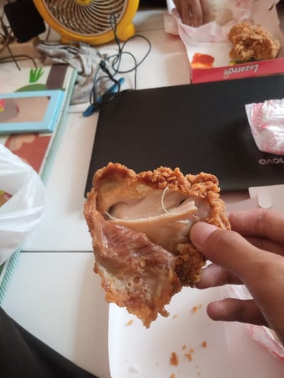The scene depicts an indoor, casual setting, presumed to be in Muka, Indonesia. A person's hand with light-toned skin is holding a piece of fried chicken, broken open to expose cooked white meat and some connective tissue. The chicken features a crispy, golden-brown breading. Below the chicken, a white fast-food container with crumbs is visible. To the left, a black fabric object, likely clothing, is partially visible.
In the midground, a black laptop mat, partially obscuring a "Lenovo" logo, rests on a white surface. To its right, an open red and white "Lazarro" branded cardboard box contains a piece of fried chicken, a portion of white rice, and a small amount of red sauce. Another hand, lighter in skin tone, is visible near this box. A crumpled white wrapper is also present.
The background shows a yellow table fan. Various items are scattered on the white surface, including a light blue-framed object (possibly a mirror), a white plastic bag, entangled black and blue cables, and a crumpled light grey cloth. A green and white striped mat is visible beneath some items. The overall environment suggests a domestic or casual eating area.