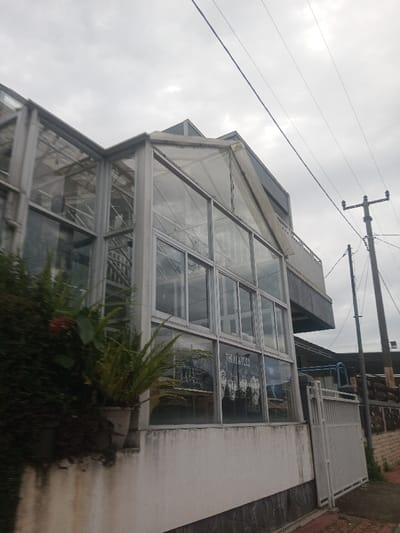 The image depicts a multi-story building located in Suddepalle, India, under an overcast sky. The structure features a prominent glass and metal-framed facade, suggestive of a conservatory or greenhouse design, particularly on its upper levels. The glass panels reflect the cloudy sky and internal structural elements. On the lower section of the building's front, a white poster featuring black silhouettes of four men and the text "THE BEATLES" is affixed to a glass panel. Below the glass, the building transitions to a white-painted wall with a dark stone or tile base. To the left, dense green foliage, including potted plants and vines, grows along the building's base, with a single red flower visible within the greenery. To the right, a white metal gate, possibly a pedestrian entrance, stands next to a low stone wall. Further to the right and receding into the background, a dark utility pole supports multiple overhead power lines that stretch across the upper right portion of the frame. An upper-level balcony with a white railing is visible on the right side of the main building, extending from a more solid, dark-colored section of the structure. No individuals are present in the scene, and no explicit actions or interactions are observed.