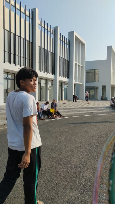 In Trosobo, Indonesia, a young man in a white t-shirt and black track pants with green stripes walks on an asphalt surface in front of a modern, multi-story building with white facades and dark-framed windows. Several people are seated on the tiered concrete steps leading up to the building, and one person is bent over near the steps. Another person in a pink headscarf stands on the steps. The sky is clear and blue, and the sun casts shadows on the ground.