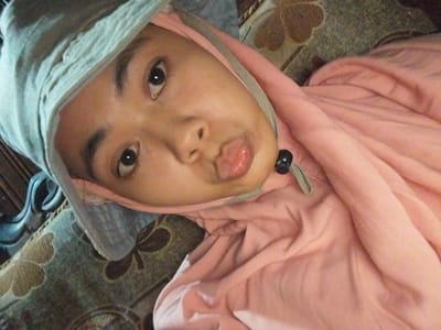 A person wearing a light gray and pink hooded garment is lying down, with their head tilted and face close to the camera. The person has dark eyes and is wearing a light gray hat that covers their head and part of their face. Their lips are slightly pursed, and they are looking directly at the camera. The garment is a soft pink color and has folds and wrinkles, suggesting it is made of a lightweight fabric. The background appears to be a patterned surface, possibly a sofa or cushion, with floral motifs in shades of brown and beige. The lighting suggests it might be daytime, but the specific time of day is unclear. The overall impression is a candid, informal shot taken indoors.