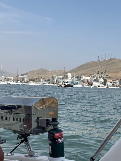 Here is a description of the image.

The image shows a view from a boat on the water, presumably near the city of Ancón, Peru, given the coastal landscape and architectural style visible in the background. The sky is clear and blue. The foreground shows a stainless steel marine-grade grill attached to the boat, with a Coleman propane tank connected. In the background, a stretch of coast is lined with white buildings, many appearing to be residential, built along the base of a large, dry, brown hill or mountain. Several sailboats are docked near the shore. The water is calm and slightly choppy. The scene is likely taken during a sunny day.