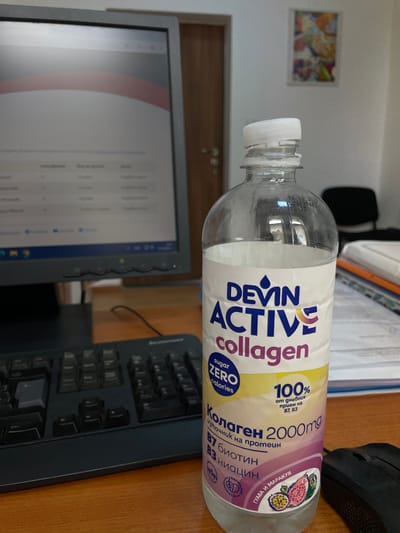 The image depicts a workspace with a bottle of Devin Active Collagen drink as the central object. The bottle is plastic, clear, and partially full. Its label is white and purple, displaying information about the drink's content, including "Collagen 2000mg," "Sugar Zero Calories," and "100% of daily intake of B7, B3."
The bottle rests horizontally on a wooden desk near a black Lenovo keyboard and a black computer mouse. A computer monitor is visible on the left side of the frame, displaying a document or spreadsheet.
The background includes a white wall, a framed artwork, and an office chair. The general setting appears to be an office environment. There is no information to associate this image with Sungurlare, Bulgaria.