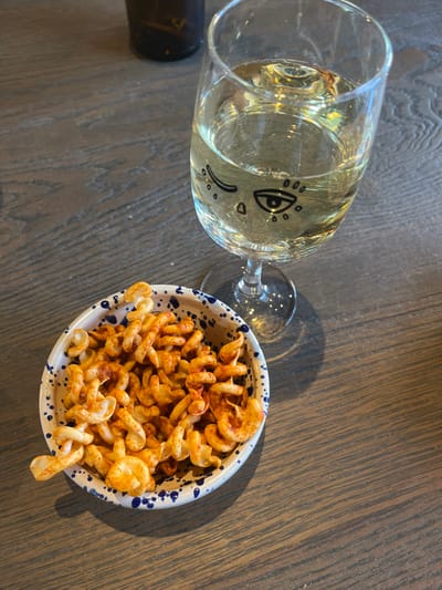This image depicts an indoor scene in Dorchester, United Kingdom, showing a white wine glass and a bowl of snacks resting on a wooden table. The clear, stemmed wine glass, positioned on the right, is approximately half-full with a pale yellow liquid consistent with white wine. Distinct black line art is drawn on the glass, featuring a winking eye on the left and an open eye with a tear-like drop below it on the right. To the lower left of the glass is a small, round ceramic bowl with a white interior and exterior, adorned with irregular blue speckles along its rim. This bowl contains numerous spiral-shaped snacks, predominantly reddish-orange from seasoning. The table surface is made of wood, exhibiting a distinct grain pattern, and appears to be a medium-dark brown hue. A dark, out-of-focus bottle is partially visible in the upper left background. Shadows from the glass and bowl are cast onto the table, indicating a light source from the upper left. No individuals are visible.