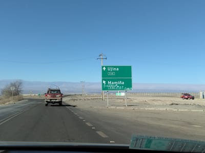 This media file depicts a highway scene in a desert environment, likely in Pozo Almonte, Chile. The main subjects are a paved road, a red pickup truck with "BHP N-01" visible on its tailgate, and a green highway signpost. The signpost has two panels. The upper panel reads "Ujina" with an arrow pointing upwards and "A-65" below it. The lower panel reads "Mamiña" with a handwritten inscription below it that appears to say "FUERZA M.O.P. CORRUP" and "A-653".
The scene appears to be set during the daytime under a clear blue sky, suggesting fair weather. The surrounding landscape is arid, characterized by sand, sparse vegetation, and distant mountains or hills. Electrical poles and wires are visible, indicating infrastructure along the highway. The presence of the pickup truck and the highway sign suggest that this is a travel or transportation context. The handwritten text on the lower sign might indicate a local sentiment or commentary. The image is taken from the perspective of someone inside a vehicle, as suggested by the dashboard and windshield visible in the foreground. No people are clearly visible, but their presence is implied by the vehicles and infrastructure.