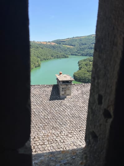 The image captures a view from a window in Castelnau-de-Mandailles, France, looking out over a reservoir and surrounding green hills. In the foreground, a tiled roof and a stone chimney are visible. The reservoir, with its calm, turquoise water, dominates the mid-ground. The background features rolling hills covered in lush green trees and some patches of dry grass, under a clear blue sky. The overall scene conveys a sense of rural tranquility and natural beauty.