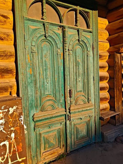 An old, turquoise-painted double door with ornate carving stands adjacent to a yellow, log-built wall. The paint on the door is chipped, revealing wood underneath, and the door itself appears weathered. The log wall has a rough texture. To the left of the door, a rusty metal surface is covered in graffiti, including white spray-painted lettering and symbols. A small wooden shelf is attached to the log wall to the right of the door. The ground in front of the door is paved with dark grey stones, and a few blades of grass are visible. The scene appears to be an exterior shot, possibly in an alley or on a street, with warm, directional lighting suggesting either morning or late afternoon sun. The location is identified as Kimry, Russia. No people are present in the image.