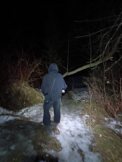 A person in dark clothing and a hooded jacket stands on a path covered in snow and patches of ice. The person is holding a flashlight. The surrounding area is wooded with bare trees and bushes. It is dark, and the person is facing away from the viewer. There is no indication in the image that shows it is in the city of Izvestkovyy, Russia.