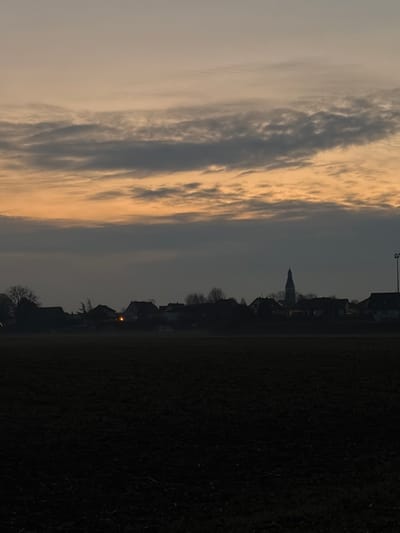An overcast sky with dramatic, layered clouds illuminated by a pale golden light dominates the left side. The right side shows a dark landscape, likely a field or open area. A silhouette of a town skyline emerges against the sky, featuring rooftops and a prominent church spire. A few bare trees are visible along the skyline, and a single light glows in a distant window. The overall atmosphere is muted and subdued.