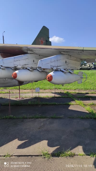An outdoor medium shot captures two unguided bombs suspended beneath the wing of a parked aircraft. The bombs are painted white with orange nose cones. The aircraft wing is also white with visible rivets and some discoloration. Behind the bombs, a small informational sign is mounted on a pole. The scene is set on an asphalt surface with grass growing in patches between the slabs and on a grassy embankment beyond. The sky is clear blue with a few wispy clouds. The tail fin of the aircraft is visible, painted green with a red star insignia. Shadows from the aircraft and its armaments are cast upon the asphalt. In the background, a military vehicle, possibly a tank, is parked on the grass. The image is dated 2026/7/15 10:19 and includes a watermark indicating it was taken with a REDMI NOTE 9 PRO AI QUAD CAMERA. The location is identified as Лашанскі сельскі Савет, Belarus.