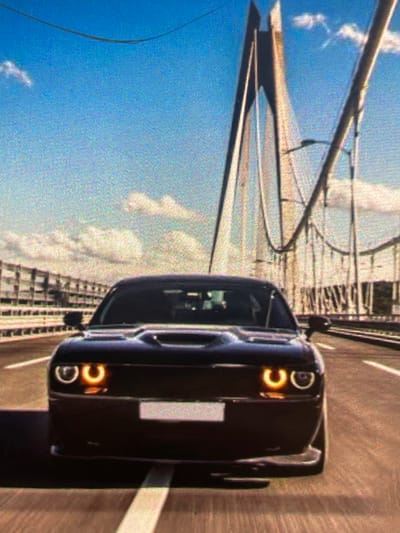 A black Dodge Challenger is shown from a low front-facing perspective, driving on a road that appears to be a bridge. The car's headlights are illuminated, emitting a yellow glow. In the background, a large suspension bridge structure with cables and pylons is visible against a blue sky with white clouds. The road is paved and has white lane markings. The overall image has a grainy texture, suggesting it may be a digital rendering or a photograph taken with a low-resolution camera. There is no indication of the location being Paczkowo, Poland.