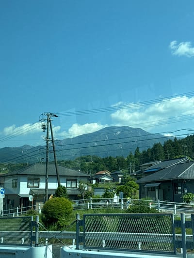 The image depicts a roadside view in Nakatsugawa, Japan. A mountain range is visible in the background, partially obscured by clouds. In the foreground, power lines stretch across the sky, running parallel to the road. Houses and trees line the road. A metal guardrail runs along the side of the road.