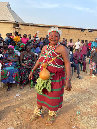 A woman stands in traditional attire, featuring a red and green striped dress, numerous colorful beaded necklaces, a gourd and greenery attached to her waist, and unique ankle accessories. A ring adorns her nose, and she wears a feathered headdress. She is surrounded by a crowd of people seated in chairs, and the setting appears to be outdoors in a rural area, with a building visible in the background.