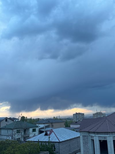 The image depicts a cityscape in Arinj, Armenia, viewed under a cloudy sky with a glimpse of sunlight on the horizon. The foreground shows rooftops of residential buildings, including a partially constructed structure with exposed brick and concrete. In the background, taller apartment buildings are visible. The sky is predominantly overcast with dark, heavy clouds.