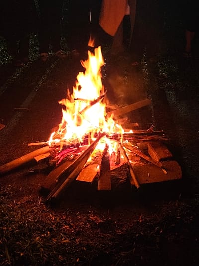 This image depicts a nighttime scene in Batulawang, Indonesia, centered on a large bonfire. The fire is constructed from numerous wooden planks and sticks, actively burning with bright orange and yellow flames that rise significantly above the fuel. The ground immediately surrounding the fire is dark earth or sparse vegetation, partially illuminated by the fire's glow, revealing scattered dry organic material. The background is predominantly dark, with faint outlines of foliage or distant trees discernible on the left. Two individuals are partially visible: one person's lower torso, legs, and a foot (possibly sandal-clad) are present on the right, standing near the fire. Another individual's arm and upper body are visible in the upper central-left, with a light-colored fabric or garment draped near them. Both individuals are positioned in close proximity to the bonfire, illuminated by its light against the otherwise dark environment. No explicit actions or interactions beyond their presence near the fire are discernible.