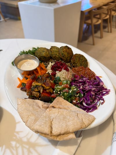 A large, white oval plate of Mediterranean cuisine is centrally displayed on a white marble or quartz-patterned tabletop. The plate contains five round, greenish-brown falafel balls resting on a bed of grains (likely quinoa), accompanied by a small metal cup of creamy white tahini sauce. To the right of the falafel, there is a portion of reddish-orange spicy sauce, and below it, a mound of hummus garnished with dark red pomegranate seeds. Further down, a vibrant green tabbouleh salad is visible, along with a distinct pile of shredded purple cabbage. Roasted mixed vegetables, including eggplant and red bell peppers, are positioned between the hummus and the shredded carrots. Two folded pieces of light brown pita bread are placed at the bottom edge of the plate.
The background, out of focus, reveals a modern, brightly lit indoor dining area, consistent with a restaurant or cafe in Las Vegas, United States. To the left, a blurred white rectangular structure, possibly a counter or partition, is visible. To the right, a long wooden dining table is partially seen with several light-colored wooden chairs, each featuring a curved backrest. The flooring appears to be a light, neutral tone. No individuals are present in the frame.