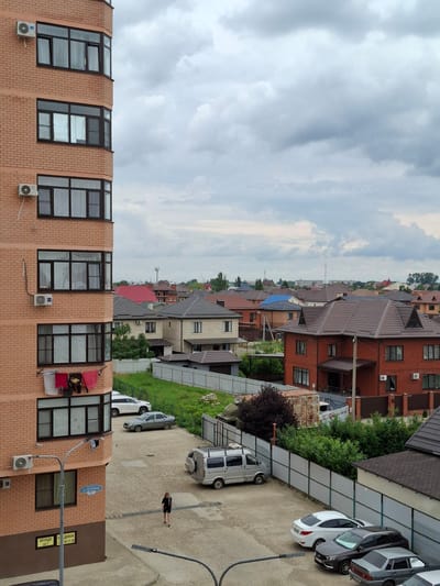 The image depicts a view of residential houses and an apartment building in Yablonovskiy, Russia. The foreground features a brick apartment building with multiple windows and air conditioning units. Clothes are drying on a line attached to the building. Below the apartment building, a paved area serves as a parking lot with several cars, including a white sedan, a dark gray station wagon, and a silver minivan. A person wearing dark clothing is walking in the parking lot. In the background, a collection of houses with various roof colors, including brown, red, and blue, are visible under a cloudy sky. A white fence runs along the perimeter of the property.