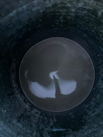 The scene, located in Cutneuheun, Indonesia, depicts a top-down view into a dark, cylindrical container with visibly textured and stained inner walls. At the bottom of the container, a circular surface of dark, reflective liquid is present. This liquid surface shows a distinct, bright, inverted crescent-shaped reflection of an overhead light source. Directly below this light reflection, a dark, elongated silhouette, interpreted as a human figure, likely the photographer holding a device, is clearly visible, extending downwards from the crescent. No other persons or objects are discernibly present within the container.
