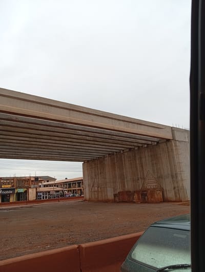 The image depicts an underpass of a large concrete bridge under construction, likely in Katambe, Nigeria. The sky is overcast and appears to be daytime, with a diffused light. The bridge structure is the dominant subject, with multiple concrete beams forming the underside of the elevated roadway. A large, weathered concrete pillar supports the bridge on the right side. On this pillar, a triangular emblem with the text "TRIA CTA" is visible. In the background, several buildings are visible, some with multiple stories and shops on the ground floor. Signs for "DHL" and "Mobile" are discernible on one of the buildings. There are some construction materials, like large concrete blocks, scattered near the pillar. A dark green car is parked in the foreground, partially obscuring the view of the road. The ground appears to be unpaved and covered in reddish-brown dirt, suggesting a dusty environment. There are no people prominently visible in the image, though some figures are present in the background near the shops. The overall scene conveys an atmosphere of ongoing construction and urban development.