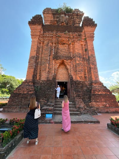 A medium shot captures a weathered, brick temple structure set against a bright blue sky with scattered white clouds. The temple, built in a style characteristic of the Cham people, features stepped tiers and a central arched entrance. Two women stand facing the temple: one in a black dress with a white tote bag on her left, and another in a long pink dress on the right. Both are viewed from behind. A person in a white robe stands at the entrance of the temple on the steps, facing outward. Lush green foliage frames the left side of the temple, while flowering plants in reddish hues line the foreground. The ground is paved with terracotta tiles. The scene appears to be set during daytime.