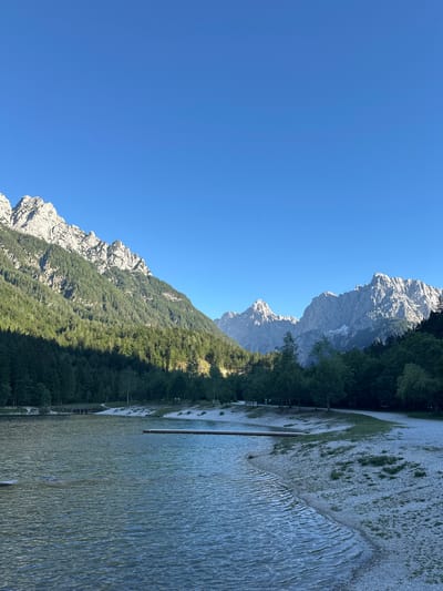 This image shows a scenic view of Lake Jasna near Kranjska Gora, Slovenia. The lake is calm with ripples on its surface, reflecting the clear blue sky. The shore is a mix of sand and pebbles, with some patches of grass. A wooden pier extends into the water from the right side of the frame. In the background, a lush green forest covers the lower slopes of majestic, rugged mountains. The mountains are rocky with sharp peaks, and some patches of snow can be seen on the higher elevations. The sky is a vibrant, cloudless blue. There are a few people visible on the sandy shore, and some trees are scattered along the waterline.