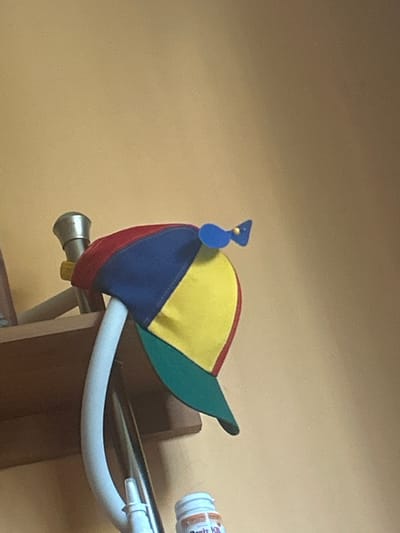 A close-up, slightly tilted, eye-level shot shows a colorful pinwheel cap perched on a white curved support. The cap, with its red, blue, yellow, and green panels, has a blue pinwheel attached to its top. To the left of the cap, a silver metallic knob with a yellow detail is visible, attached to a white horizontal bar. The background is a plain, light yellow wall. The scene is indoors, on a shelf, likely in a residential setting. No specific location in Tomaszów Mazowiecki, Poland, can be determined from the image.