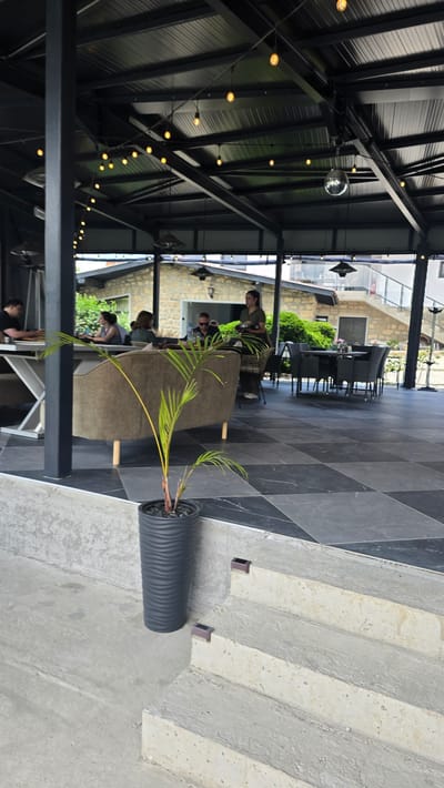 The image depicts an outdoor seating area of a restaurant or cafe, likely located in Asparuhovo, Bulgaria, as suggested by the prompt. The space is covered by a dark, sloped roof supported by black metal beams, from which decorative string lights are hung. A disco ball is visible near the center of the roof.
In the foreground, a potted palm plant stands on a tiered concrete step. The floor of the seating area is tiled in a checkerboard pattern of black and grey marble. To the left, a group of people are seated at tables, with one person appearing to be a waiter or server carrying a tray. A large, upholstered sofa is positioned in the mid-ground. In the background, another seating area with tables and chairs is visible, along with a stone building and lush green vegetation. The overall impression is of a modern, open-air dining establishment.