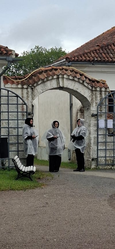 Three individuals are standing under a stone archway in front of a building with a tiled roof, under an overcast sky. They are all wearing dark clothing and transparent hooded ponchos. The person on the left is holding a small, rectangular object. The person in the middle is also holding a similar object. The person on the right is holding their hands together in front of them. To the left of the archway is a black metal gate and a white and black slatted bench. The ground is paved with gravel and there is green grass to the left. The building appears to be white with weathered stone detailing around the archway. This scene is located at the entrance of the Kuressaare Episcopal Castle in Kuressaare, Estonia.