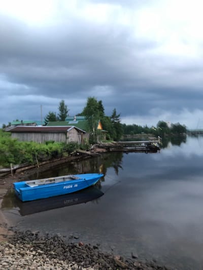 The image depicts a serene waterside scene in Svirstroy, Russia, under an overcast sky with dramatic, dark clouds. In the foreground, a bright blue boat with white trim is partially beached on a rocky shore, its reflection visible in the calm water. The boat is inscribed with Cyrillic lettering, "P5626 ЛЯ". The shore is composed of dark rocks and sparse vegetation. Behind the boat and to the left, a rustic wooden structure, possibly a shed or boathouse with a corrugated metal roof, is situated amongst lush green bushes and trees. Further back, several residential buildings with green roofs are visible, suggesting a small village or settlement. A wooden pier extends into the water from the right side of the image, with what appears to be a smaller boat or structure moored to it. The water is dark and reflective, mirroring the sky and the surrounding landscape. The overall atmosphere is tranquil and somewhat somber, characteristic of a cloudy day in a northern climate.