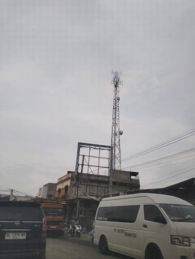 The image shows a street scene in Blangpeuria, Indonesia, under an overcast sky. A tall, lattice cellular tower dominates the center of the frame, with several antenna dishes attached at different levels. To the left of the tower is a partially constructed or decaying structure with a metal frame and what appears to be a billboard. In the foreground, several vehicles are visible. A black SUV with a license plate reading "BL 1379 NH" is on the left. To its right, an orange truck is parked. A white van, identified by the text "PT. PUTRI EMBARTICA" on its side, is in the middle ground, facing right. A person on a motorcycle is also visible in the middle ground, moving away from the viewer. The buildings in the background appear to be a mix of older, multi-story structures with ornate architectural details and more utilitarian construction. Electrical wires are strung across the scene, contributing to the urban feel. The overall impression is that of a busy, albeit somewhat dilapidated, commercial or residential area.