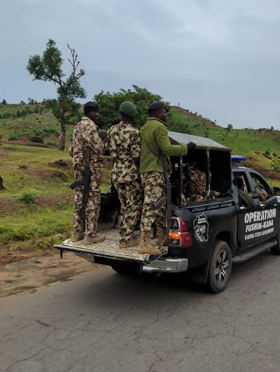 Three uniformed soldiers stand in the bed of a black pickup truck labeled "OPERATION FUSHIN-KADA, KADUNA STATE GOVERNMENT." The soldiers wear camouflage uniforms and helmets. Two hold rifles. The truck is on a paved road beside a grassy hillside with a tree. The location is suggested as Gidan Waya, Nigeria.