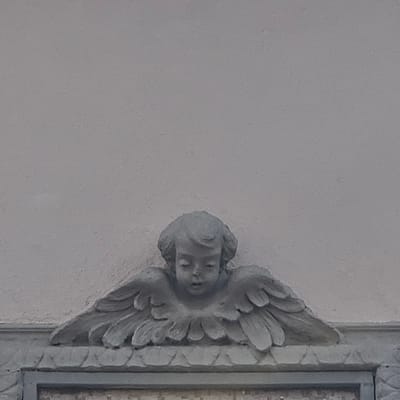 The image shows a close-up, eye-level view of a stone carving of an angel's head and wings, set against a textured, pale gray wall. The angel carving is monochromatic and appears weathered, with details of its curly hair, closed eyes, and small nose clearly visible. Its wings are spread outwards and downwards, layered and stylized. Below the angel carving is a decorative stone ledge with a repeating triangular pattern. The overall impression is one of architectural ornamentation, likely part of a building's facade in Calcinate, Italy.