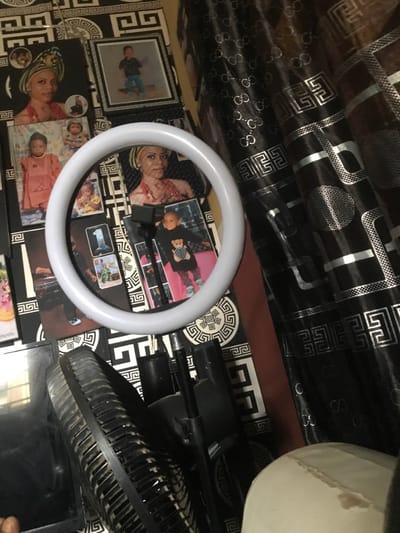 This image presents an indoor scene located in Oba-Ile, Nigeria. The foreground is dominated by a white circular ring light, mounted on a black stand. Behind the ring light, a wall is densely covered with photographs and a black and white geometric Greek key patterned wallpaper. Numerous unframed and framed photographs depict young children in various poses and attire, including one child in a traditional garment and another in a black top with a teddy bear design. A prominent adult female, wearing a headwrap and traditional jewelry, is featured in multiple photographs. To the right, several rolls of black material with silver "GC" brand-like logos and repeating geometric patterns are stacked vertically. In the lower left quadrant, the dusty grill of a black electric fan is partially visible. Additional black stand components are present beneath the ring light's mounting point. A small portion of a light-colored object, possibly upholstery, is visible in the lower right corner.