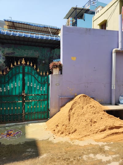 The scene depicts an outdoor residential area in Pedda Kammavaripalli, India, under clear daylight. A large, conical pile of sandy-brown aggregate rests on a concrete and dirt ground surface in the foreground, positioned against a building wall.
To the left, a two-story building features a weathered, dark teal facade with extensive peeling paint and an upper window secured by a wrought-iron grill. Its roof structure consists of blue-trimmed corrugated metal with an additional blue railing visible above. Directly in front of this building is a closed, dark teal double gate constructed of ornate wrought iron, adorned with golden finials and decorative swan-shaped motifs.
To the right of the gate, a building wall exhibits a pale lavender exterior with visible cracks and areas of paint loss. A white PVC downspout runs vertically along this wall, connecting to a horizontal pipe near the base of the sand pile. A white potted plant with dark reddish-green foliage sits on a concrete ledge protruding from this wall, adjacent to the gate.
On the ground directly in front of the gate, a colorful Rangoli design is present, featuring purple and white outlines filled with yellow and orange floral elements. Portions of the ground appear damp, and strong shadows are cast across the lower foreground. Above the structures, a clear blue sky is visible. No individuals, actions, or interactions are observed.