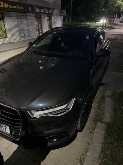 An Audi A6 Avant is parked on a street at night in Orhei, Moldova. The car is dark gray and features a license plate with the number "87". In the background, a building with a sign that reads "to market" and "Bucuria" is visible. The street is paved with concrete and there is a curb with grass on the side. The scene appears to be illuminated by streetlights and the car's headlights.